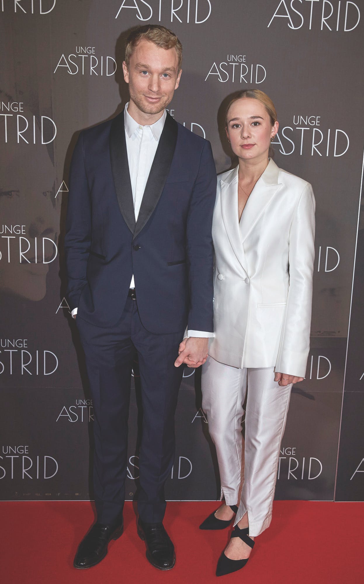 Alba August and Björn Gustafsson at the premiere of 'Beyond Astrid' holding hands with each other, both wearing pant suit. He wears blue and she wears white with black heels
